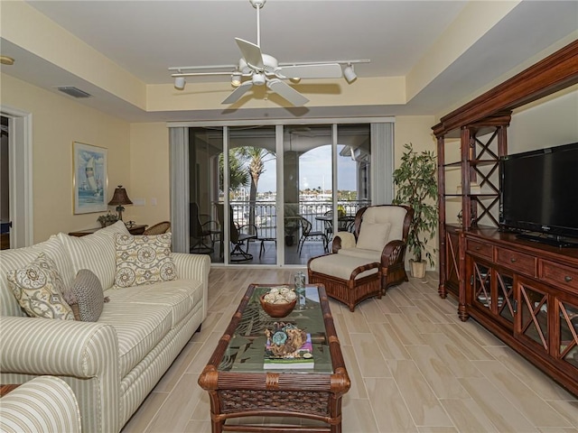 living room with ceiling fan, rail lighting, a raised ceiling, and light wood-type flooring