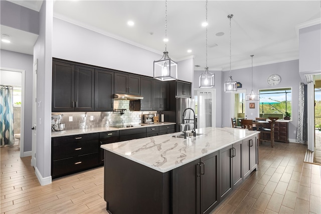 kitchen with light stone counters, hanging light fixtures, an island with sink, ornamental molding, and light hardwood / wood-style flooring