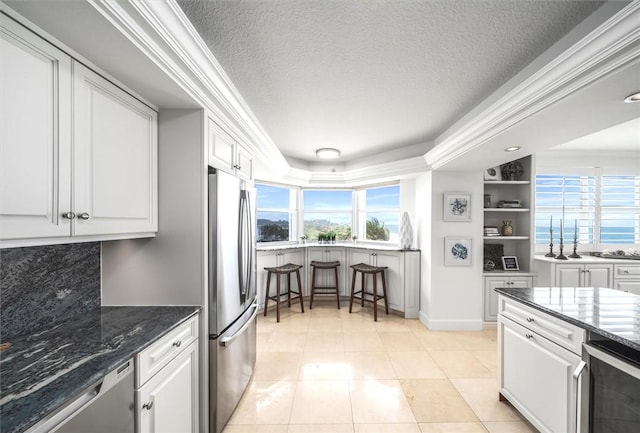 kitchen featuring stainless steel appliances, crown molding, light tile patterned floors, dark stone countertops, and white cabinets