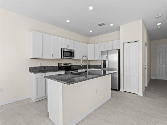 kitchen with dark stone counters, stainless steel appliances, sink, white cabinetry, and an island with sink