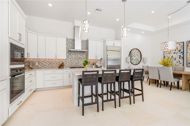 kitchen with built in appliances, a center island with sink, wall chimney range hood, and white cabinets