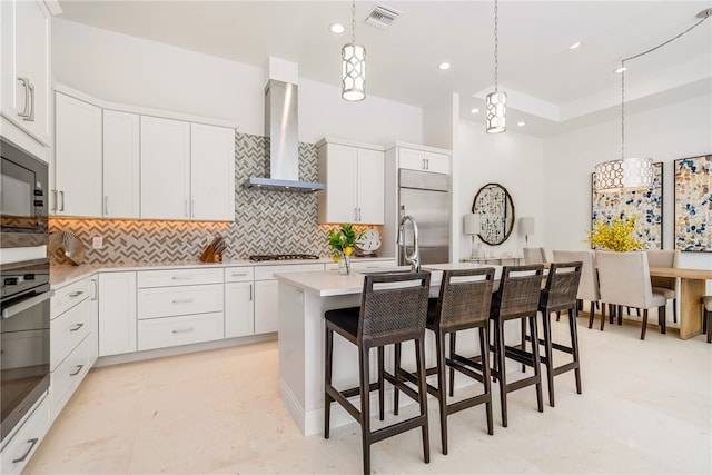 kitchen with built in appliances, a center island with sink, white cabinetry, decorative light fixtures, and wall chimney range hood