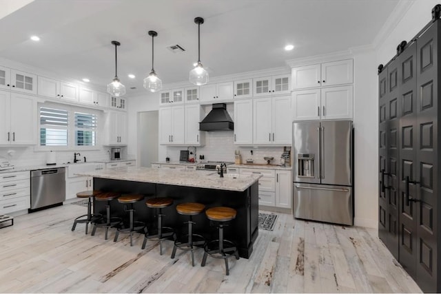 kitchen featuring wall chimney range hood, appliances with stainless steel finishes, white cabinetry, a center island with sink, and a barn door