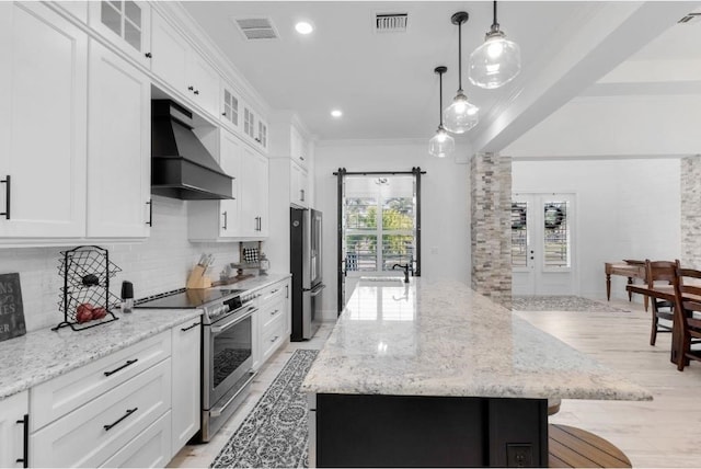 kitchen featuring premium range hood, a kitchen island with sink, hanging light fixtures, white cabinetry, and stainless steel appliances