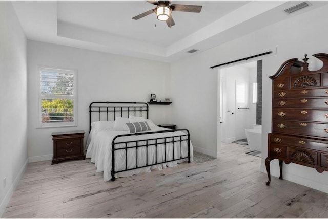 bedroom with ceiling fan, ensuite bath, a tray ceiling, and light wood-type flooring