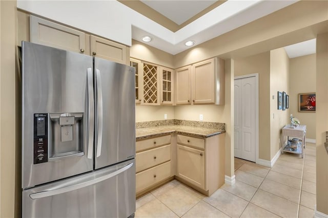 kitchen with stainless steel fridge with ice dispenser, glass insert cabinets, light stone counters, light brown cabinetry, and light tile patterned flooring