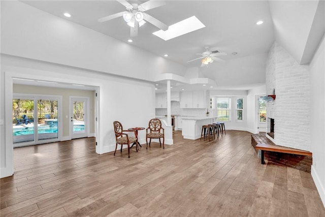 living area featuring a skylight, high vaulted ceiling, a brick fireplace, light hardwood / wood-style flooring, and ceiling fan