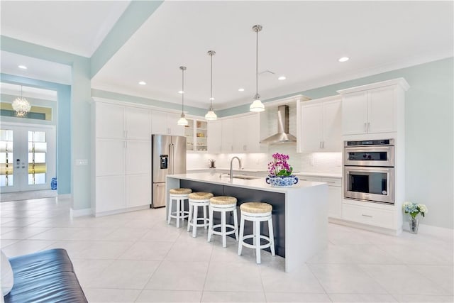 kitchen featuring white cabinetry, hanging light fixtures, wall chimney range hood, an island with sink, and appliances with stainless steel finishes