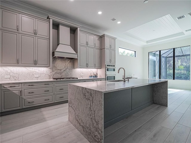 kitchen with tasteful backsplash, a center island with sink, light wood-type flooring, appliances with stainless steel finishes, and custom range hood