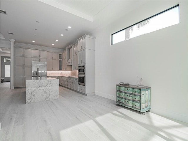 kitchen featuring gray cabinets, appliances with stainless steel finishes, a kitchen island with sink, tasteful backsplash, and light stone countertops