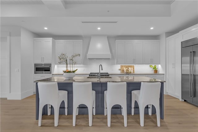 kitchen featuring an island with sink, custom range hood, white cabinets, and appliances with stainless steel finishes