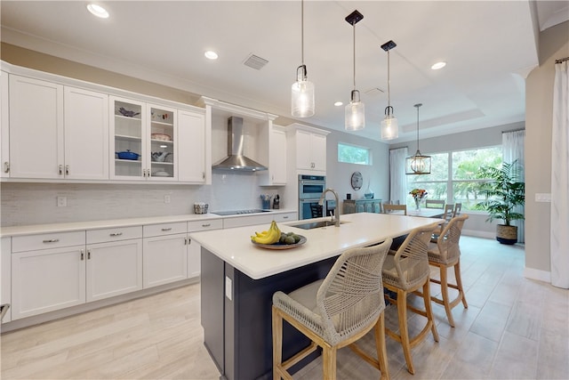 kitchen featuring hanging light fixtures, wall chimney range hood, white cabinetry, and a center island with sink