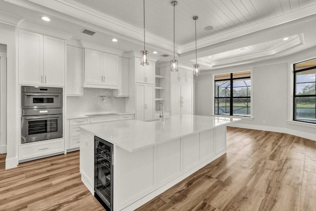 kitchen with white cabinetry, decorative light fixtures, a tray ceiling, an island with sink, and beverage cooler