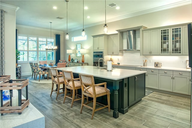 kitchen with a center island with sink, ornamental molding, wall chimney exhaust hood, light hardwood / wood-style flooring, and double oven