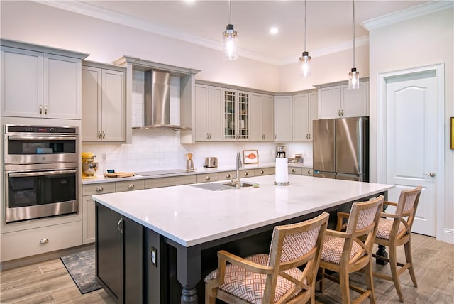 kitchen featuring sink, appliances with stainless steel finishes, wall chimney exhaust hood, a kitchen island with sink, and gray cabinetry