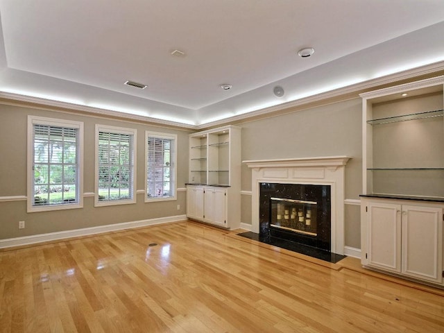 unfurnished living room with a tray ceiling, light wood finished floors, visible vents, a high end fireplace, and baseboards