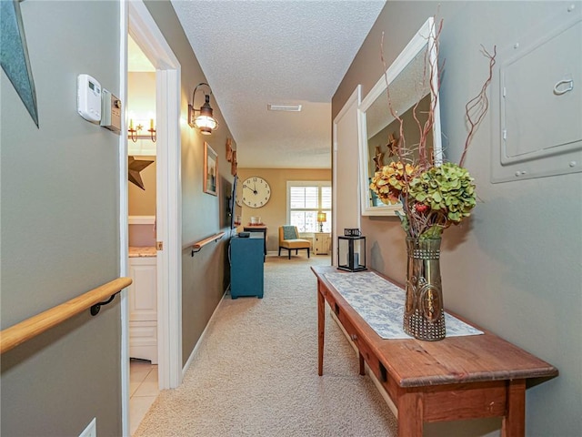 hall with a textured ceiling, light colored carpet, visible vents, baseboards, and electric panel