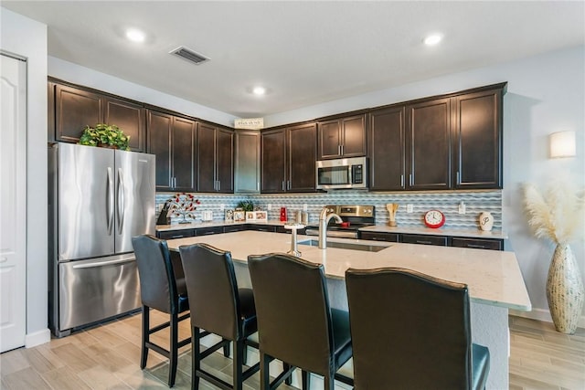 kitchen with decorative backsplash, dark brown cabinetry, stainless steel appliances, and a center island with sink