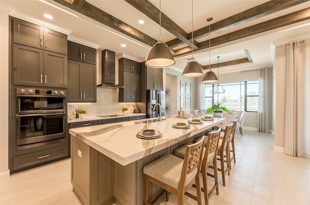 kitchen with tasteful backsplash, light stone counters, decorative light fixtures, wall chimney exhaust hood, and a kitchen island with sink