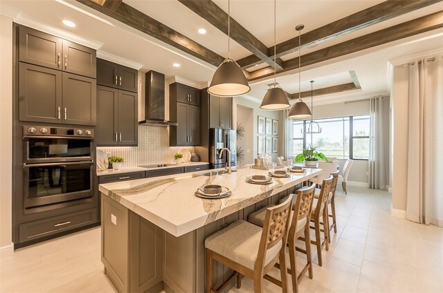 kitchen with tasteful backsplash, light stone counters, decorative light fixtures, wall chimney exhaust hood, and a kitchen island with sink