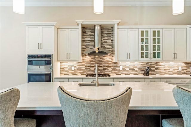 kitchen featuring white cabinets, wall chimney range hood, crown molding, hanging light fixtures, and tasteful backsplash