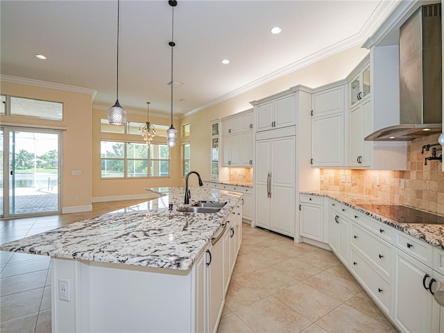 kitchen featuring white cabinets, sink, an island with sink, wall chimney range hood, and decorative light fixtures