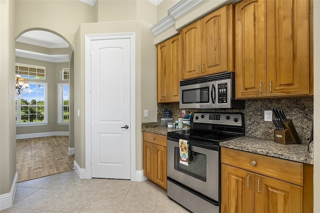 kitchen with crown molding, light tile patterned floors, dark stone countertops, appliances with stainless steel finishes, and decorative backsplash