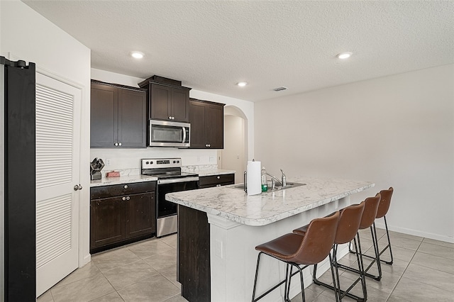 kitchen with dark brown cabinetry, stainless steel appliances, an island with sink, a textured ceiling, and a breakfast bar area