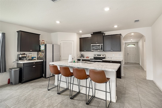 kitchen with dark brown cabinetry, sink, stainless steel appliances, a textured ceiling, and a kitchen island with sink