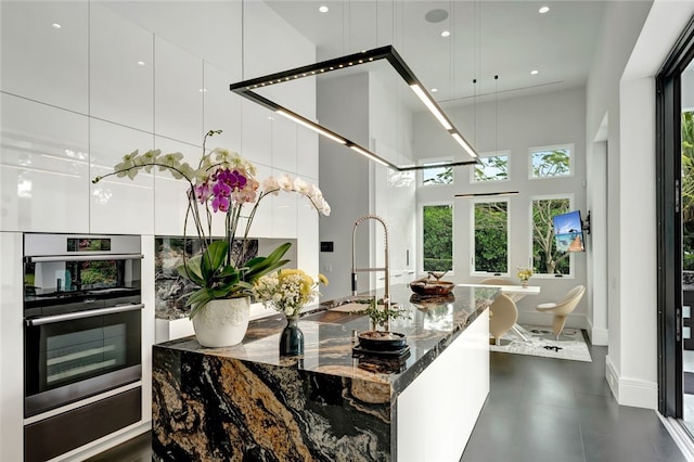 kitchen with white cabinetry, a towering ceiling, dark stone counters, double oven, and sink