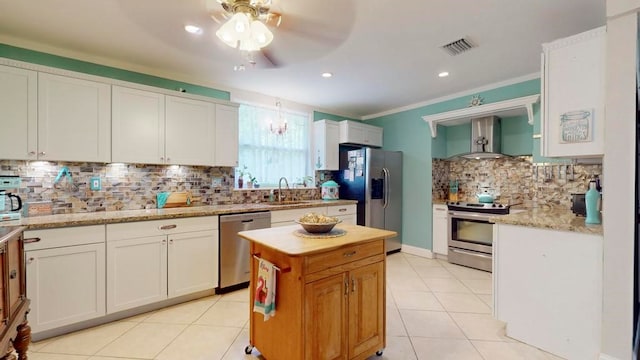 kitchen featuring crown molding, white cabinetry, appliances with stainless steel finishes, wall chimney range hood, and light stone countertops