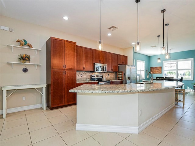 kitchen featuring tasteful backsplash, appliances with stainless steel finishes, decorative light fixtures, light stone countertops, and a kitchen island with sink