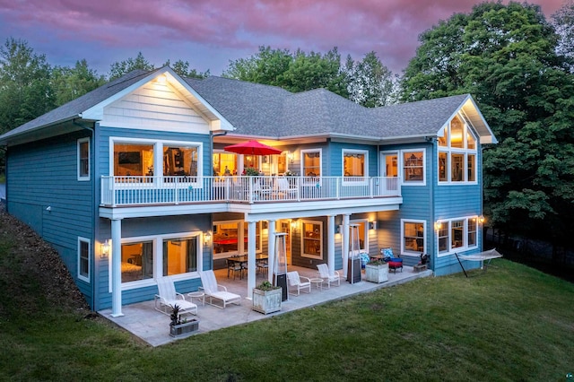 back house at dusk with a balcony, a patio, and a yard