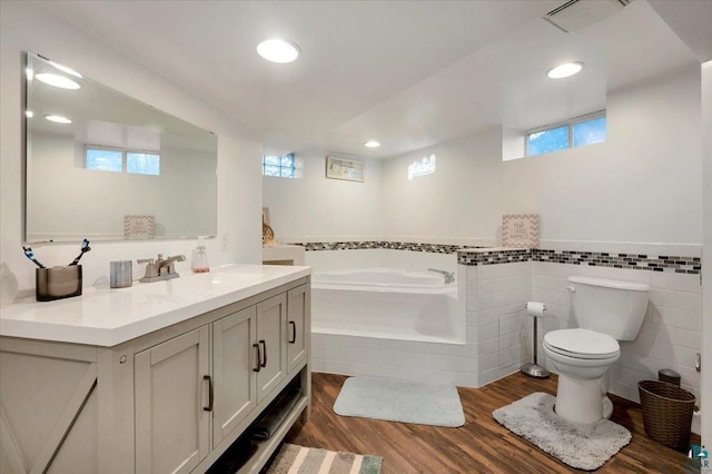 bathroom with tile walls, tiled bath, wood-type flooring, vanity, and toilet