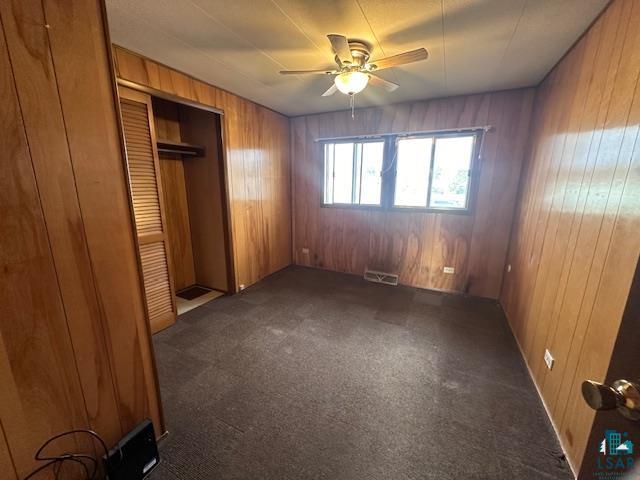 unfurnished bedroom featuring a closet, wood walls, ceiling fan, and dark carpet