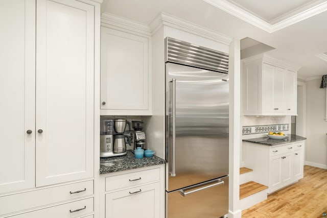 kitchen with light wood-type flooring, dark stone counters, stainless steel built in refrigerator, crown molding, and white cabinets