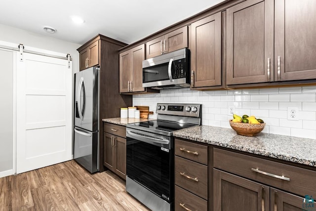 kitchen with dark brown cabinetry, light stone countertops, a barn door, light hardwood / wood-style flooring, and appliances with stainless steel finishes