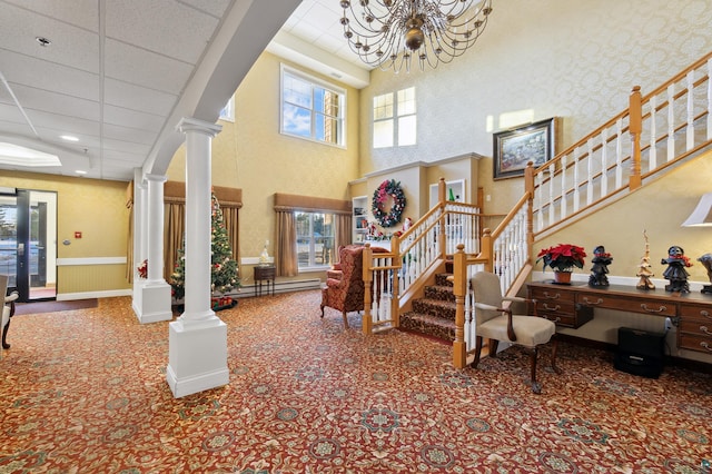 carpeted foyer entrance with a towering ceiling, ornate columns, a wealth of natural light, a drop ceiling, and a notable chandelier