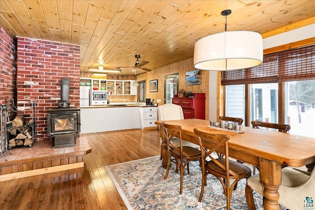 dining area with wood ceiling, wood walls, light wood-type flooring, ceiling fan, and a wood stove