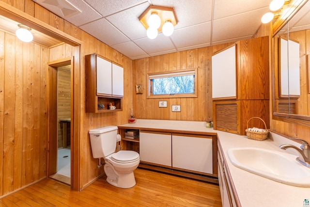 bathroom featuring wooden walls, vanity, wood-type flooring, a drop ceiling, and toilet