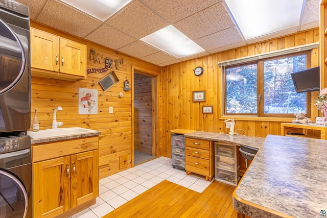 kitchen featuring stacked washing maching and dryer, wooden walls, sink, light hardwood / wood-style floors, and a drop ceiling