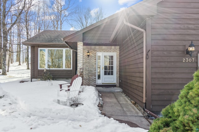 snow covered property entrance featuring brick siding and roof with shingles