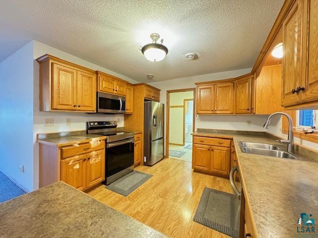 kitchen with a textured ceiling, stainless steel appliances, a sink, light wood-type flooring, and brown cabinetry