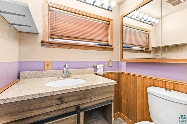 half bathroom with wooden walls, visible vents, toilet, a wainscoted wall, and vanity