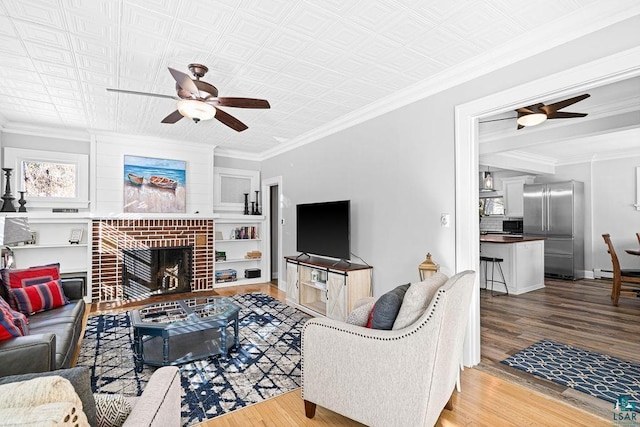 living area with baseboards, a ceiling fan, wood finished floors, crown molding, and a brick fireplace