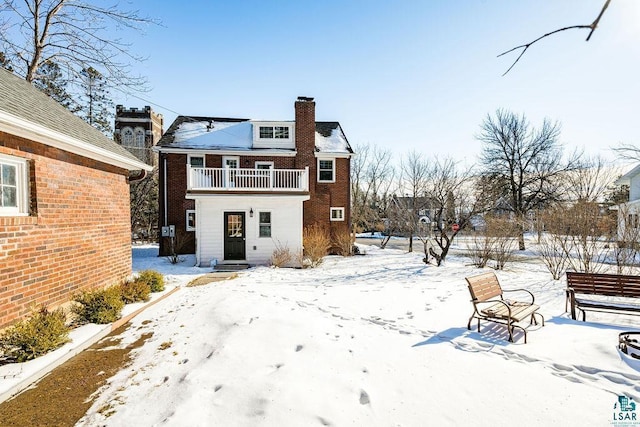 snow covered rear of property featuring brick siding, a chimney, and a balcony