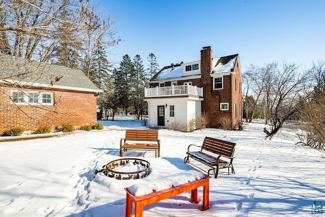 snow covered back of property featuring a balcony, an outdoor fire pit, a chimney, and brick siding