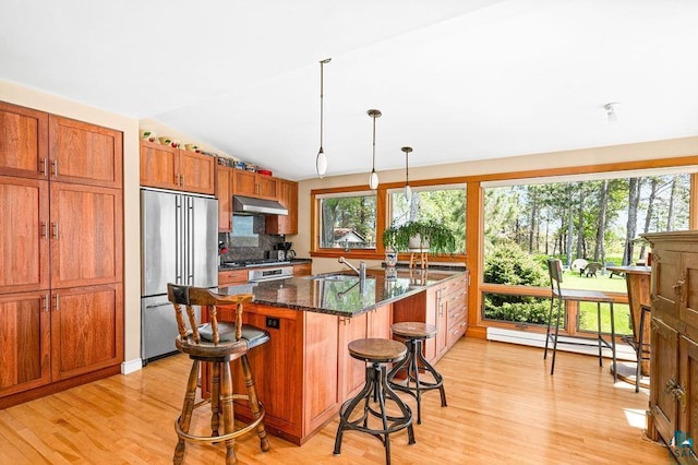 kitchen with stainless steel appliances, baseboard heating, brown cabinetry, a sink, and under cabinet range hood