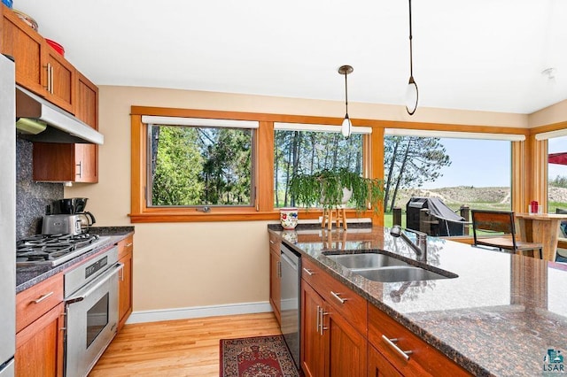 kitchen featuring appliances with stainless steel finishes, brown cabinetry, dark stone counters, a sink, and light wood-type flooring