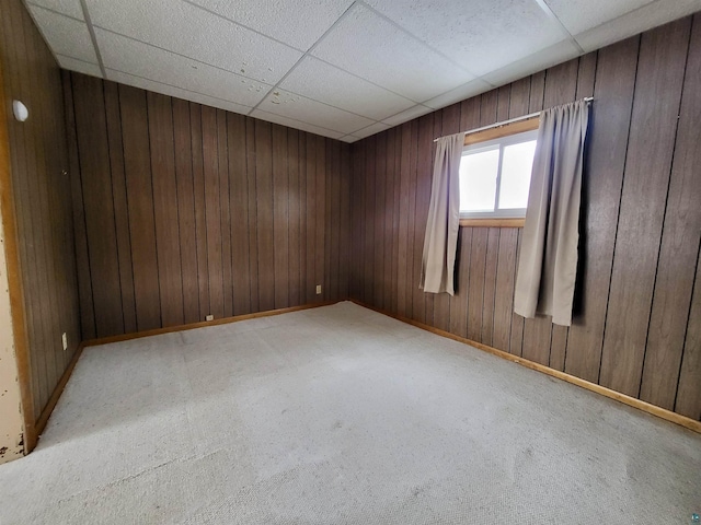 empty room with a paneled ceiling, wood walls, light carpet, and baseboards